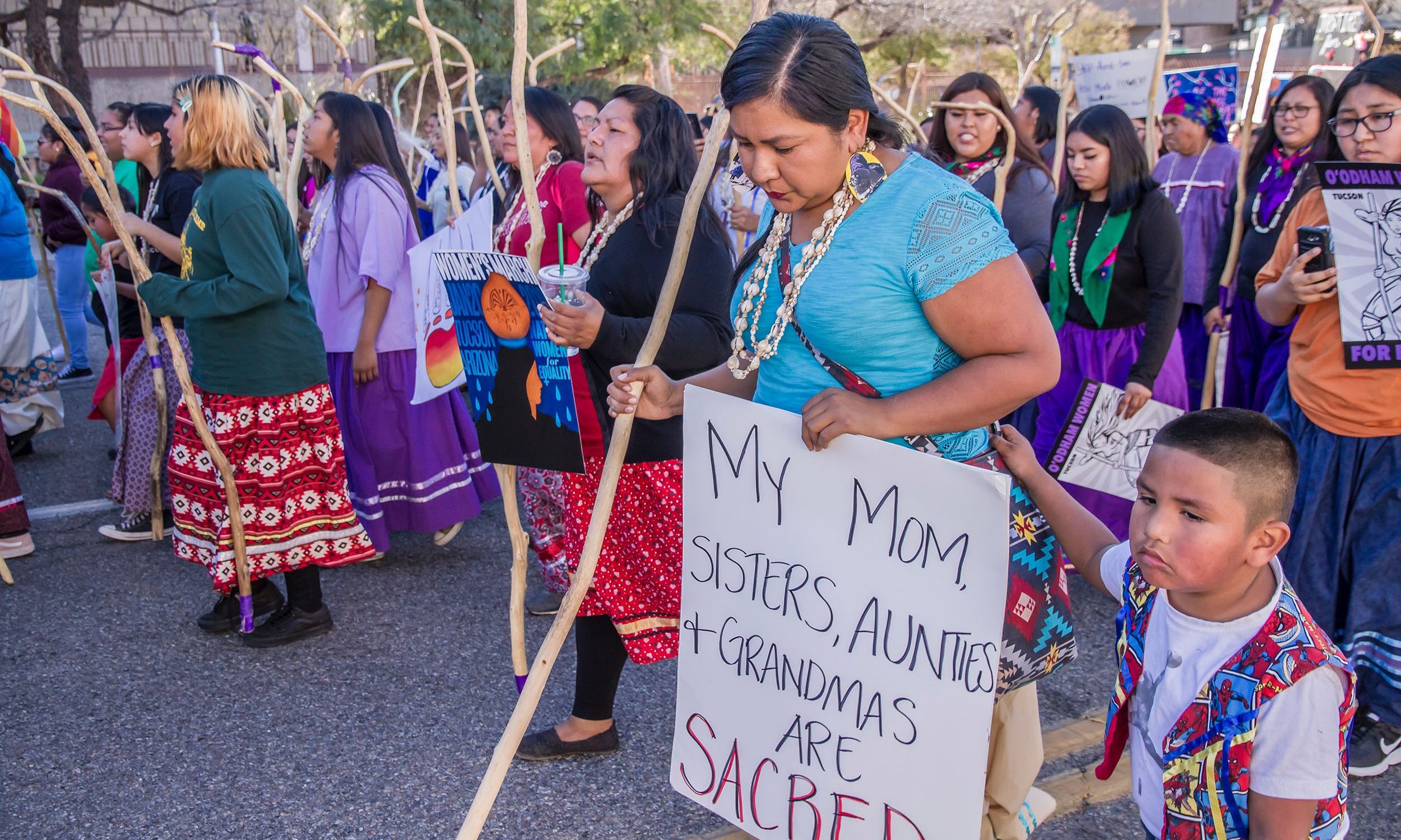 woman in protest holding sign that reads 'My mom, sisters, aunties + grandmas are sacred' (link to file)