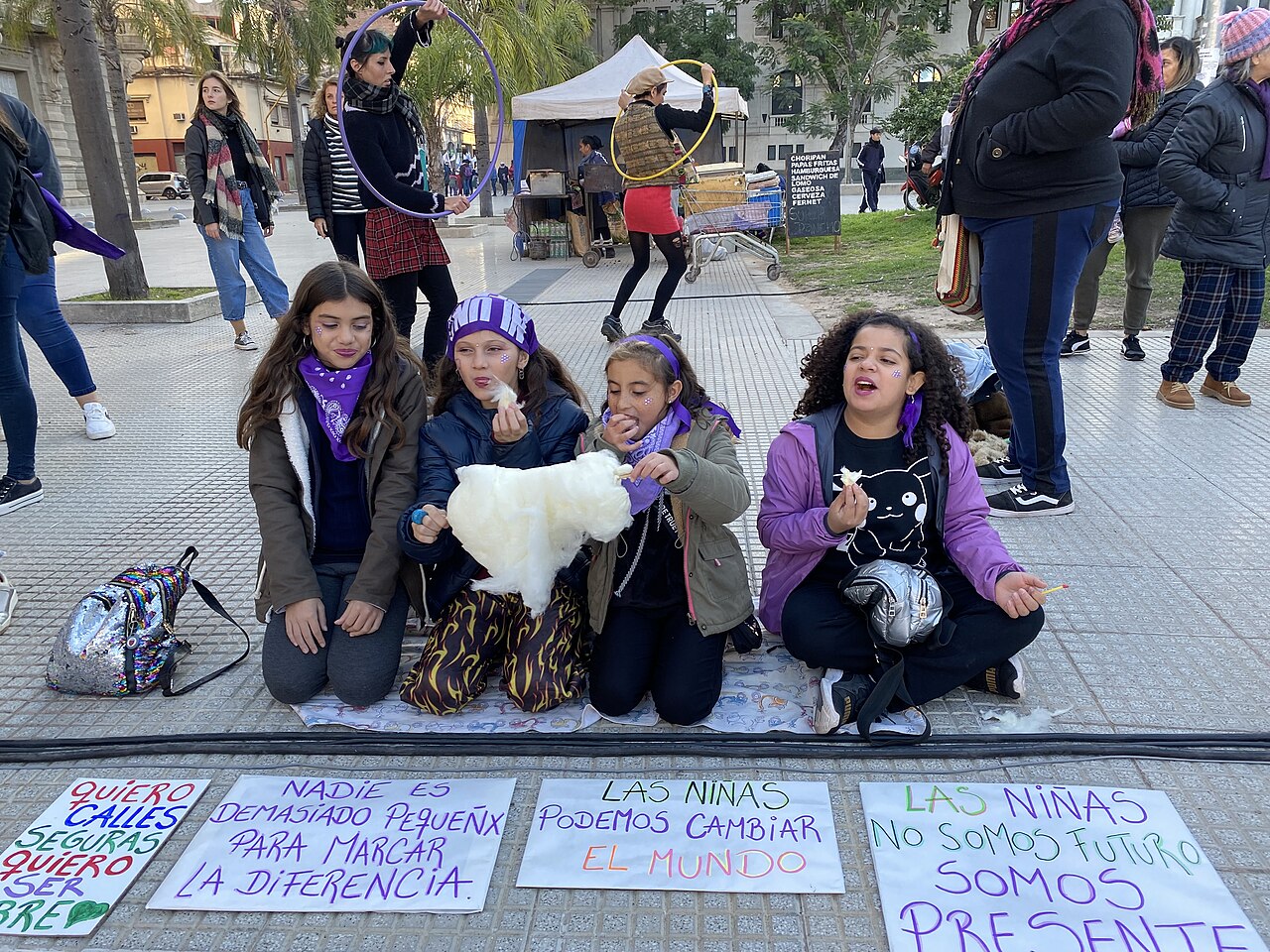 Four young women wearing purple, kneeling on the sidewalk in front of Spanish-language protest signs