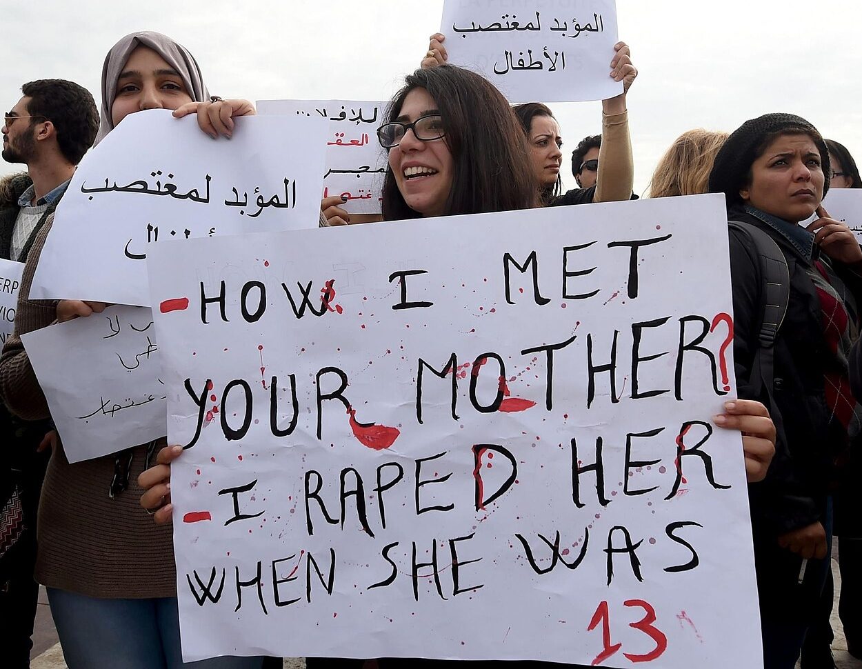 A group of protesters with signs in Arabic and English. The front sign reads “HOW I MET YOUR MOTHER? I RAPED HER WHEN SHE WAS 13”