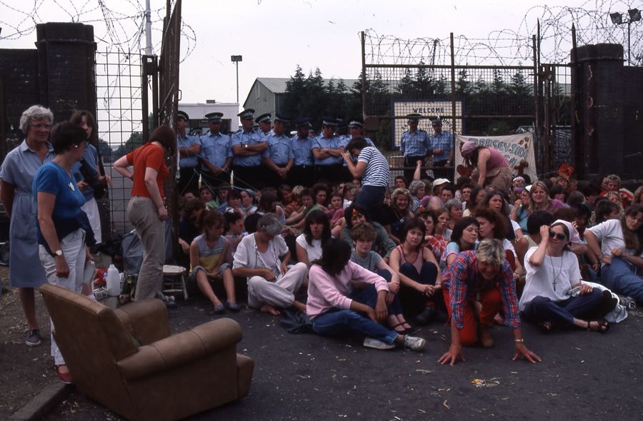 A large group of women protestors sit in front of a barbed wire fence with an open gate blocked by uniformed police officers